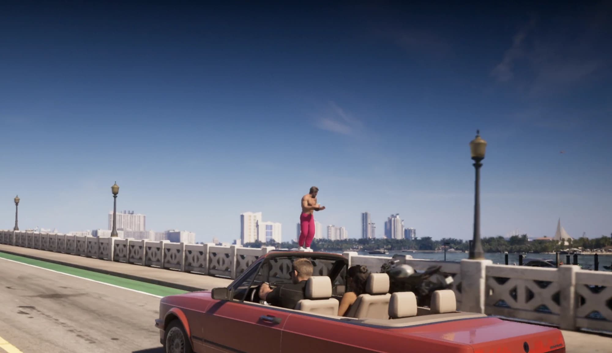 Un homme torse nu en pantalon rose debout sur le parapet d’un pont, pendant que Jason et Lucia passent en cabriolet rouge